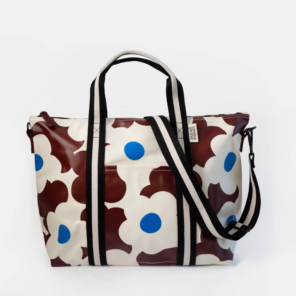 A weekend bag featuring a bold floral pattern in dark burgundy, cream, and bright blue, with black and cream striped handles and a matching shoulder strap, set against a plain white background.
