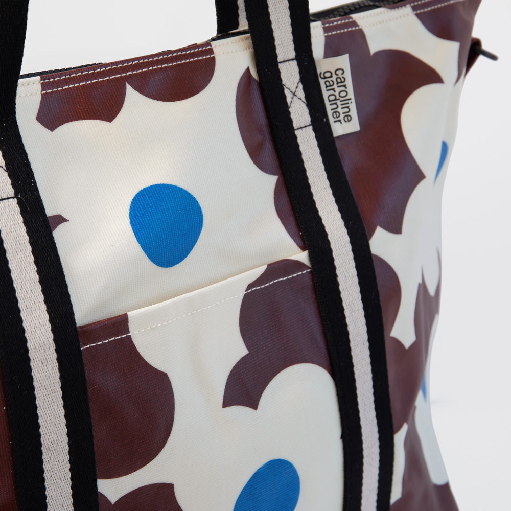 A close-up of a tote bag with a bold floral pattern in brown and blue on an off-white background, featuring black and white striped handles and a visible 'Caroline Gardiner' label.