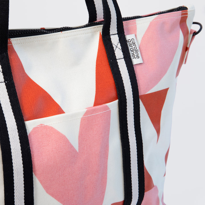 A close-up of a bag with a white base, large pink and orange abstract shapes, black and white striped handles, and a visible Gardine Garage brand tag.
