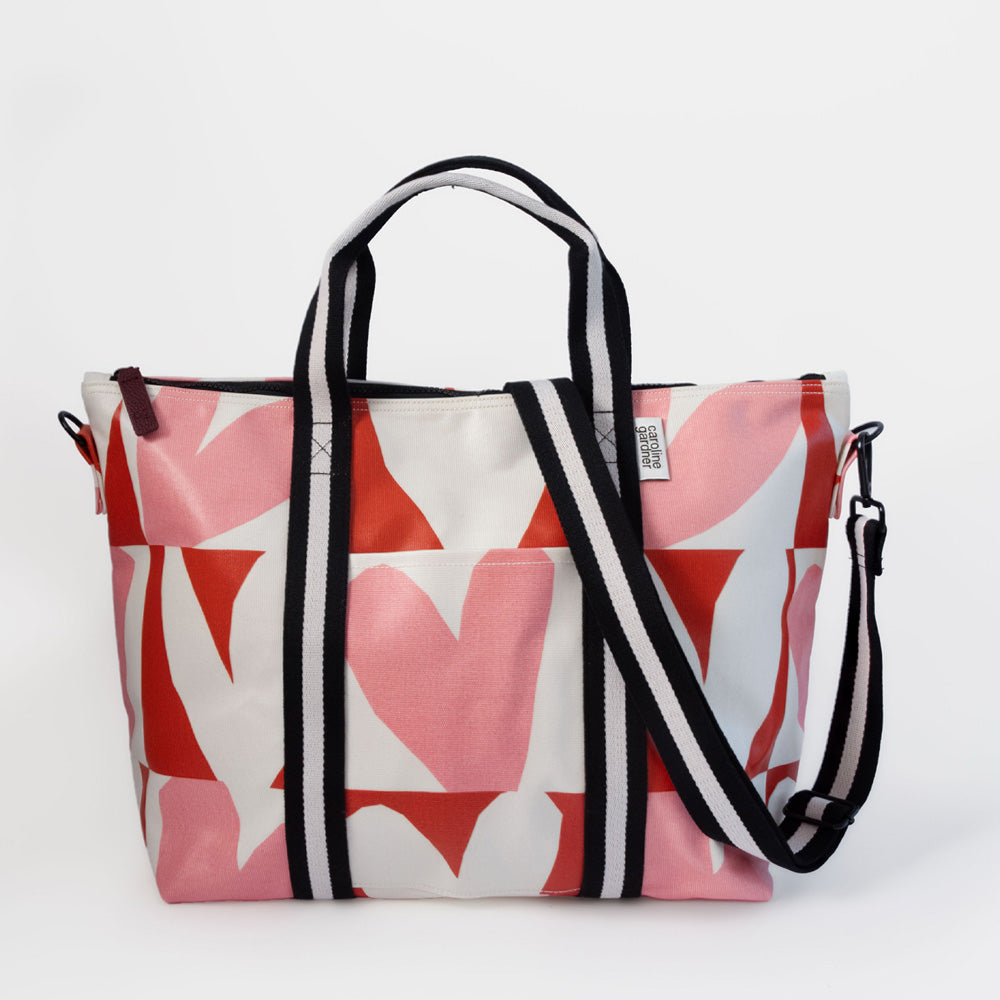 A weekend bag featuring a bold pink, red, and white geometric pattern, with black and white striped handles and a matching shoulder strap, set against a plain white background.