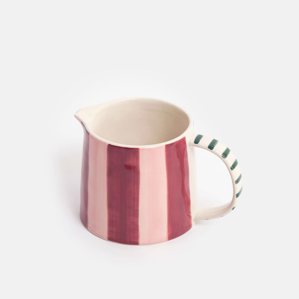 A ceramic jug with vertical pink and deep red stripes on the body, a cream-colored interior, and a handle decorated with green diagonal stripes, set against a plain light gray background.