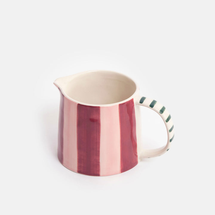 A ceramic jug with vertical pink and deep red stripes on the body, a cream-colored interior, and a handle decorated with green diagonal stripes, set against a plain light gray background.