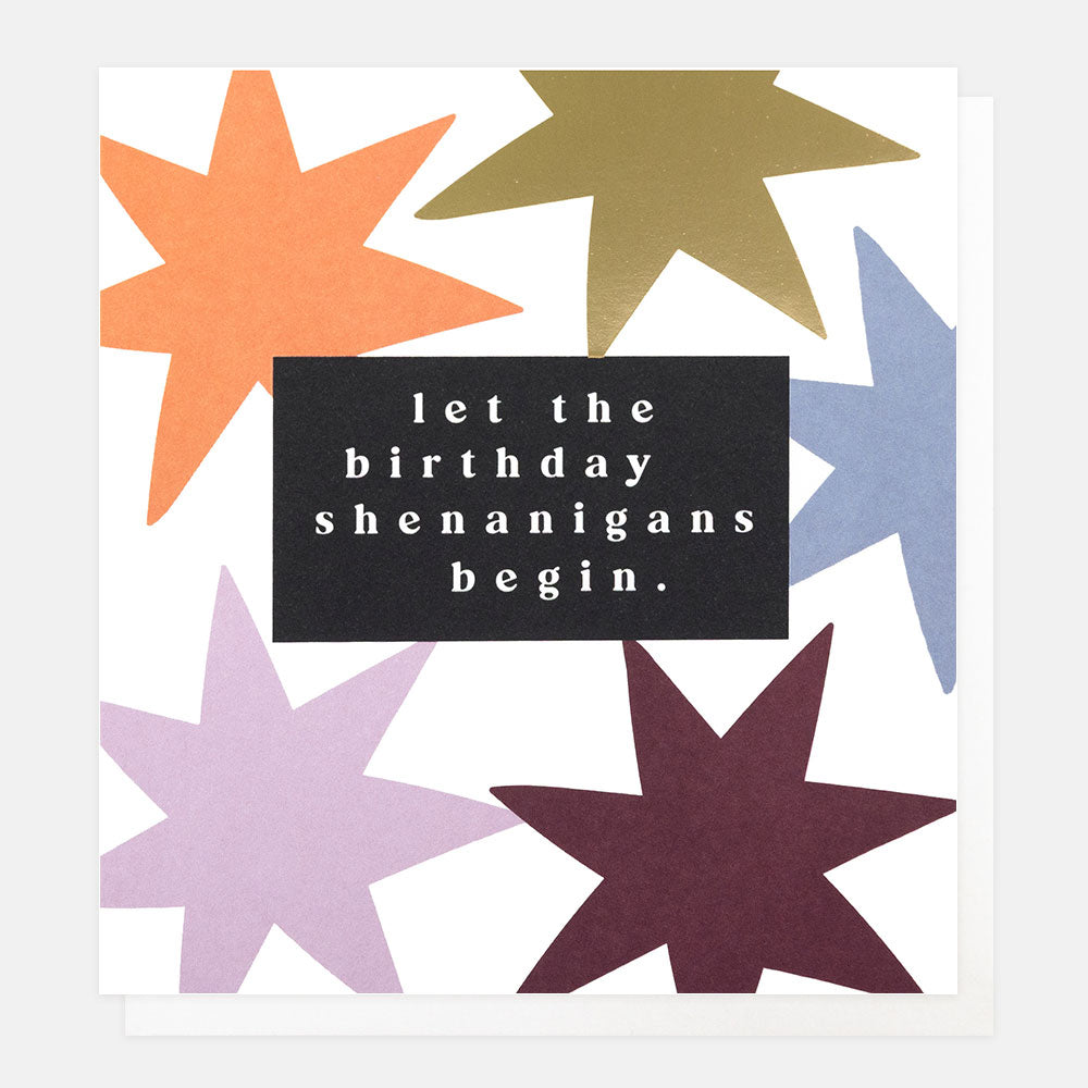 A white greetings card with large orange, lilac, blue, plum and gold foil stars and the text 'let the birthday shenanigans begin.' in white embossed lettering on a black panel, shown with a white envelope behind it.