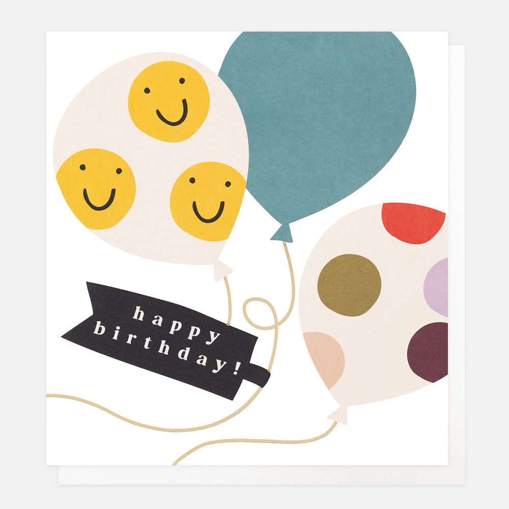 A white greetings card with balloons covered in yellow smiley faces and colourful dots, gold foil strings and the text 'happy birthday!' in white lettering on a black banner, shown with a white envelope behind it.