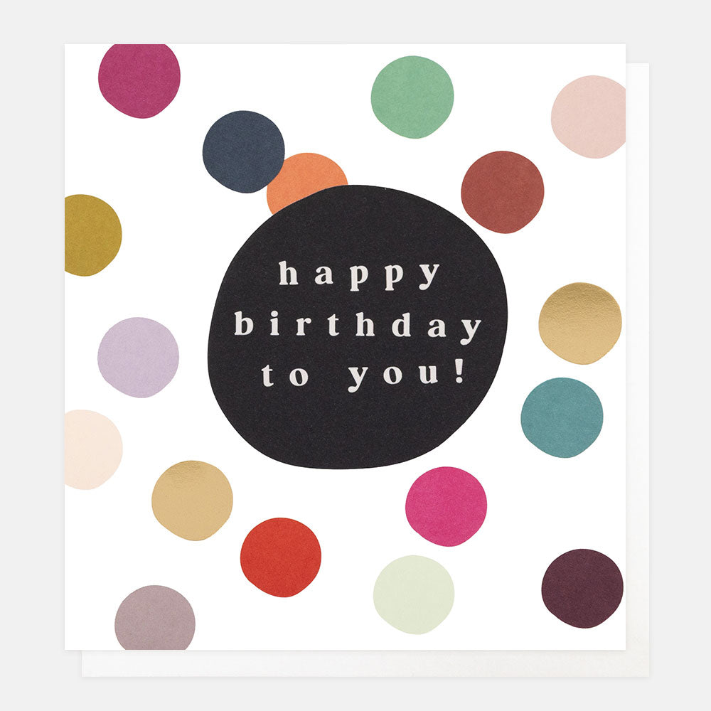 A white greetings card scattered with colourful and gold foil dots around a black bubble with the text 'happy birthday to you!' in white lettering, shown with a white envelope behind it.