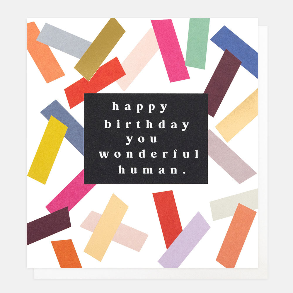 A white greetings card with colourful and gold foil confetti strips and the text 'happy birthday you wonderful human.' in white lettering on a black panel, shown with a white envelope behind it.