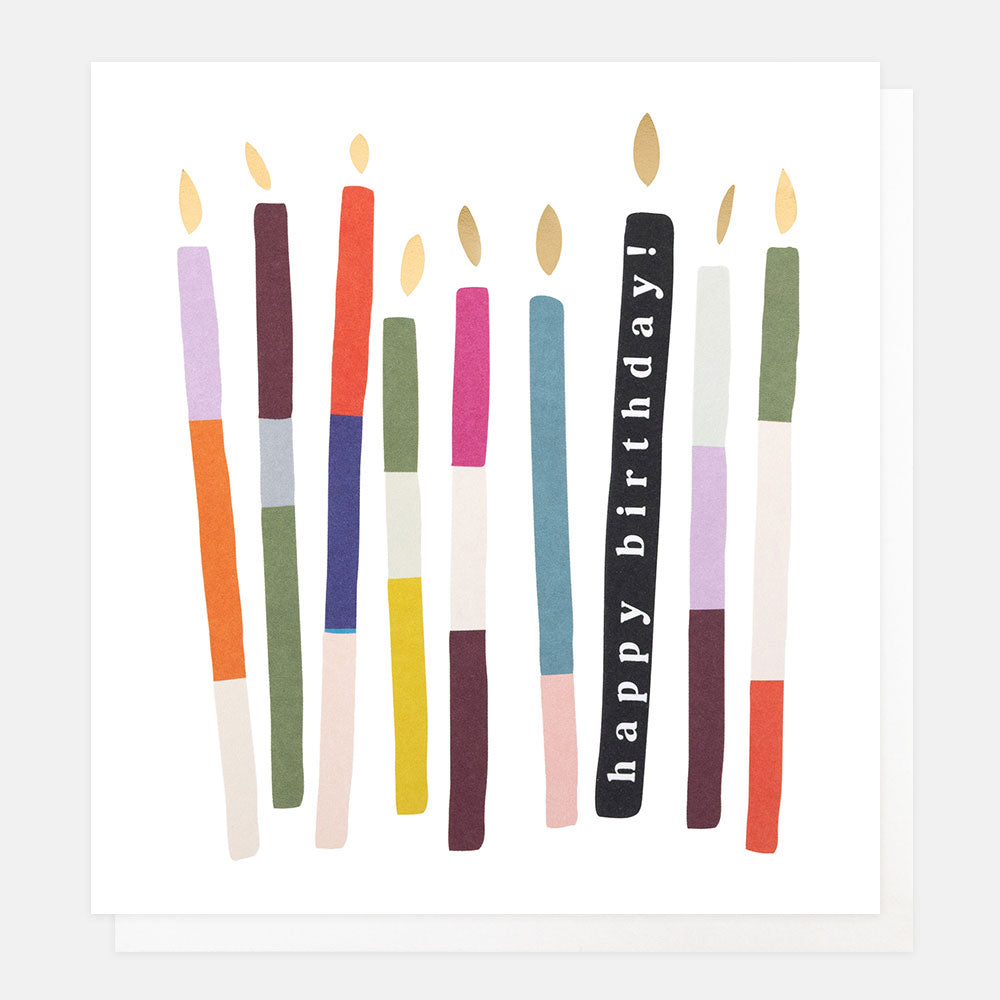 A white greetings card with a row of multi-coloured striped candles with gold foil flames, one black candle reading 'happy birthday!' in white lettering, shown with a white envelope behind it.