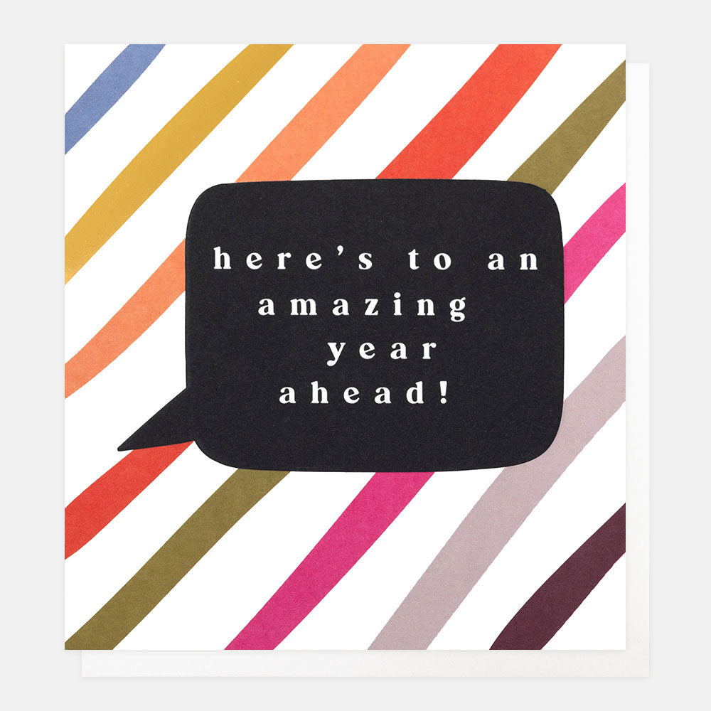 A greetings card with multi-coloured and gold foil diagonal stripes and a black speech bubble with the text 'here's to an amazing year ahead!' in white lettering, shown with a white envelope behind it.