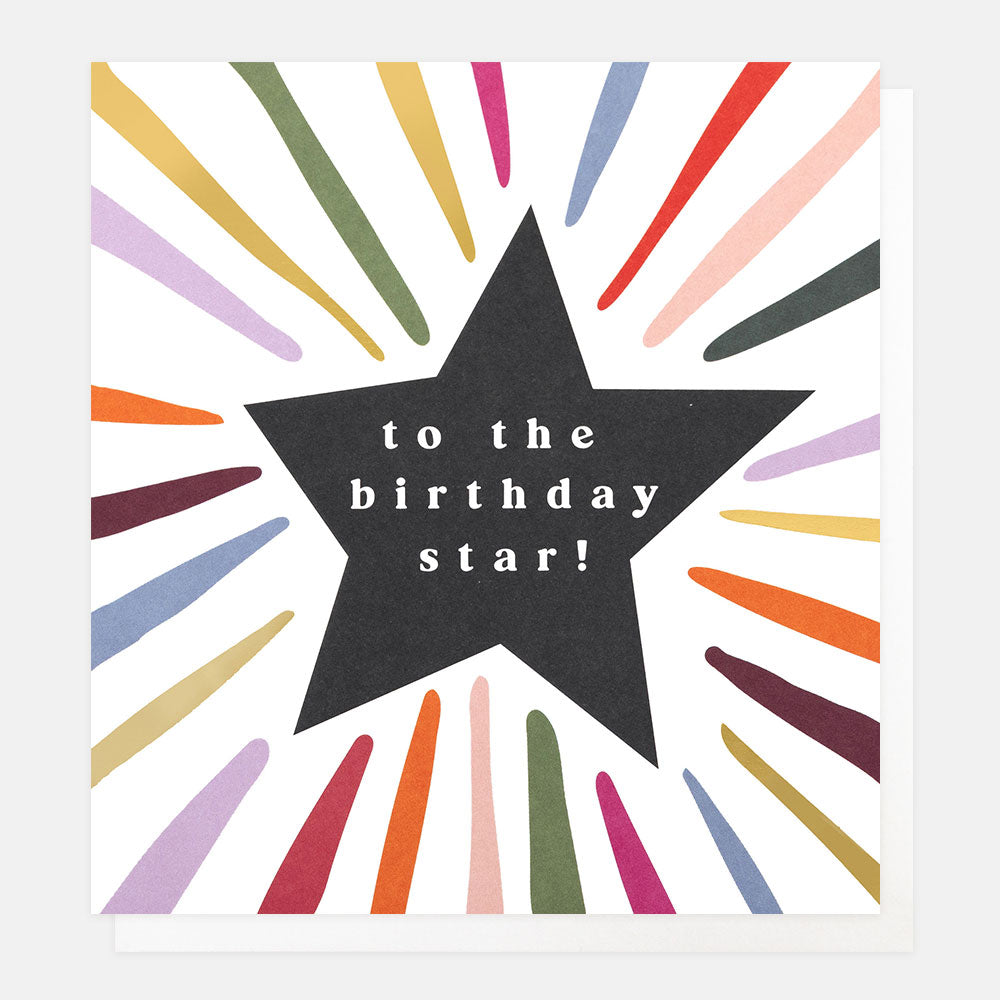 A white greetings card with a black star surrounded by multi-coloured and gold foil rays and the text 'to the birthday star!' in white lettering, shown with a white envelope behind it.