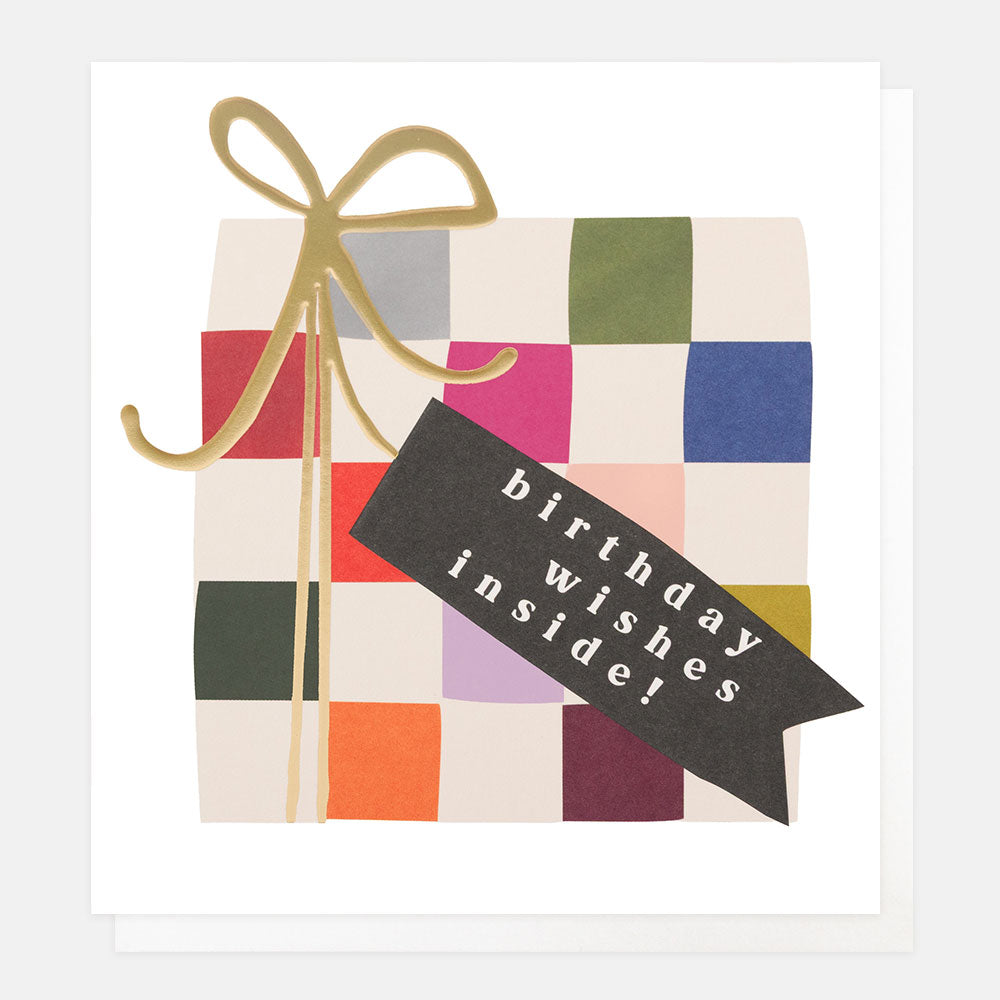 A white greetings card with a multi-coloured check present tied with a gold foil bow and a black tag reading 'birthday wishes inside!' in white lettering, shown with a white envelope behind it.