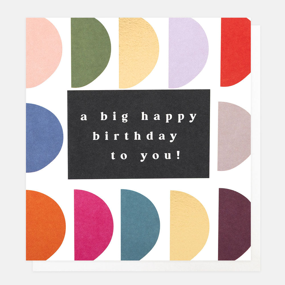 A greetings card with rows of multi-coloured and gold foil semi-circles and the text 'a big happy birthday to you!' in white lettering on a black panel, shown with a white envelope behind it.