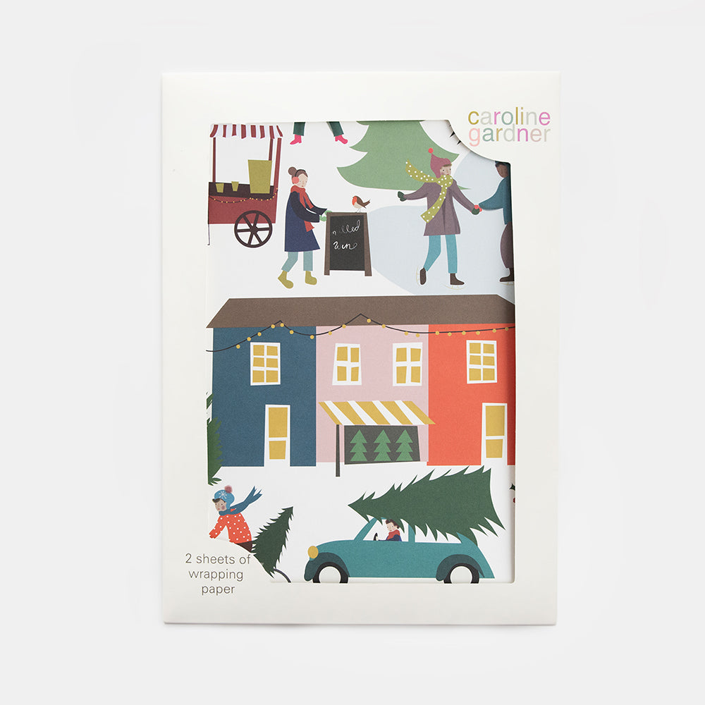 A package of Caroline Gardner Christmas wrapping paper featuring a colorful winter village illustration with houses, people, a Christmas tree, and a car, shown on a light gray background.