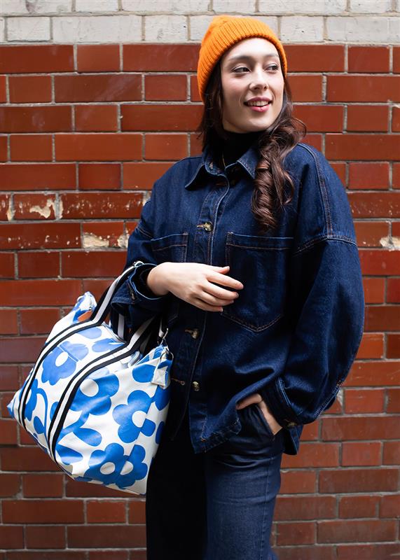 woman in blue denim jumpsuit standing in front of red brick wall holding a blue floral weekend bag