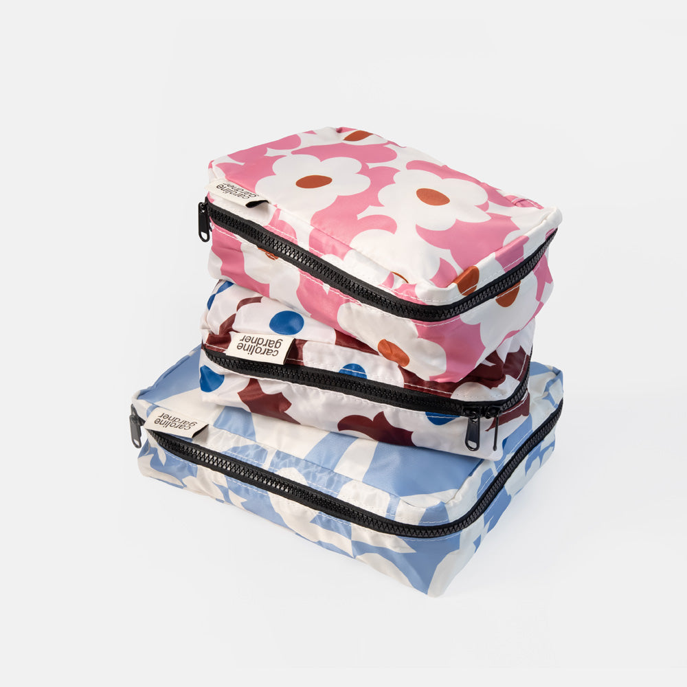 Three stackable zippered travel packing cubes with black zippers, featuring a pink and white daisy pattern on top, a blue, white, and brown pattern in the middle, and a blue and white checkered pattern on the bottom. A small white tag on each cube reads 'Packing Cubes by Amanda'.