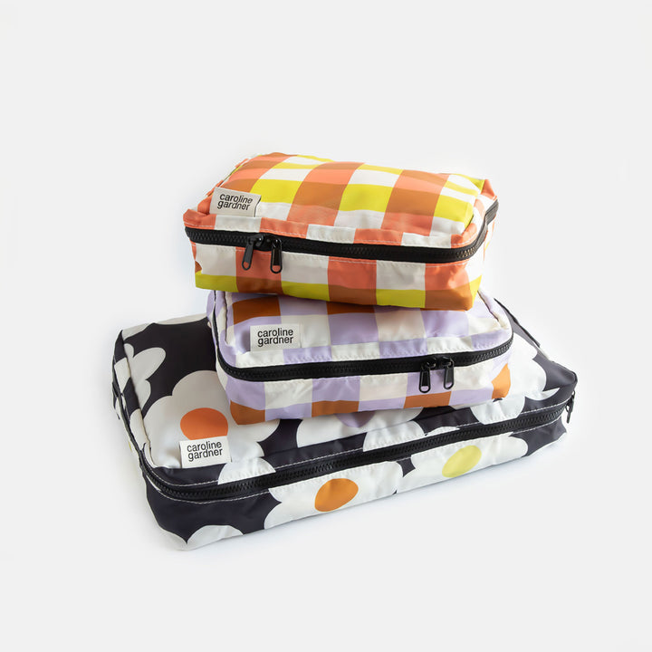 set of 3 packing cubes. 1 large in a cream flowers on a charcoal background design and 2 smaller cubes in lilac and yellow/orange check designs