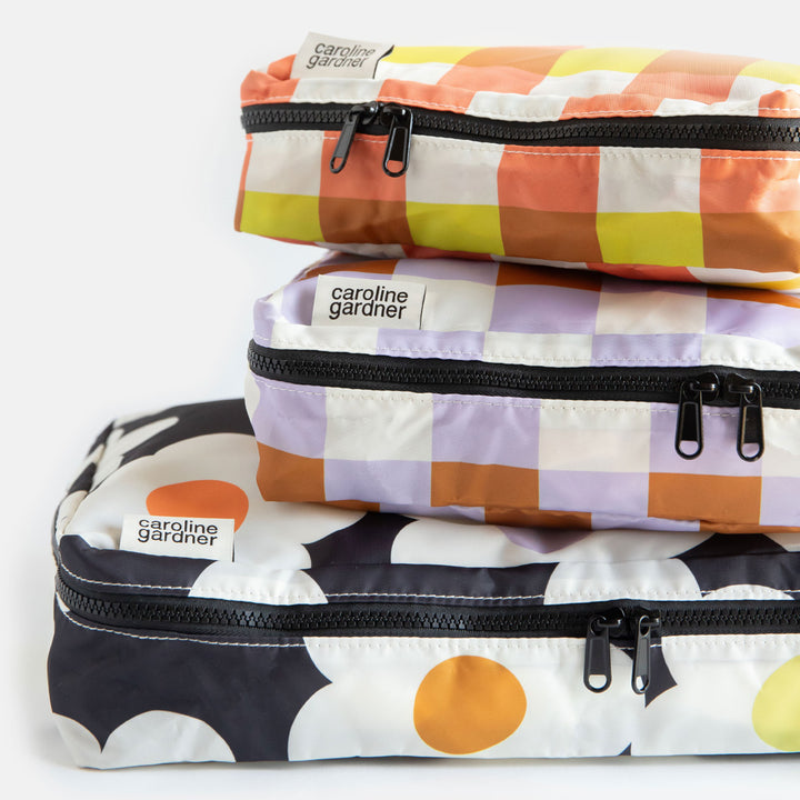 set of 3 packing cubes. 1 large in a cream flowers on a charcoal background design and 2 smaller cubes in lilac and yellow/orange check designs