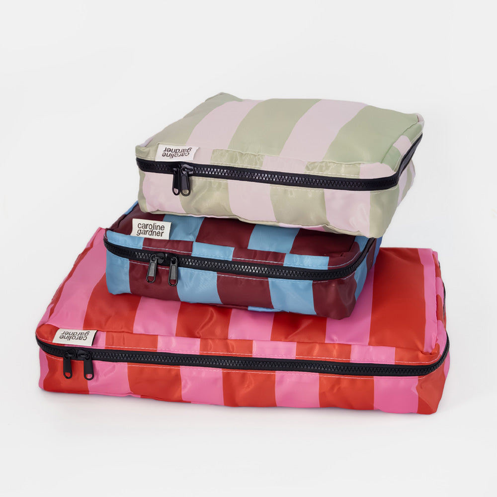 Three stacked zippered packing cubes in different pastel and bright stripe patterns on a clean white background. The top cube has light green and pink stripes, the middle has blue and burgundy stripes, and the bottom has pink and coral stripes.