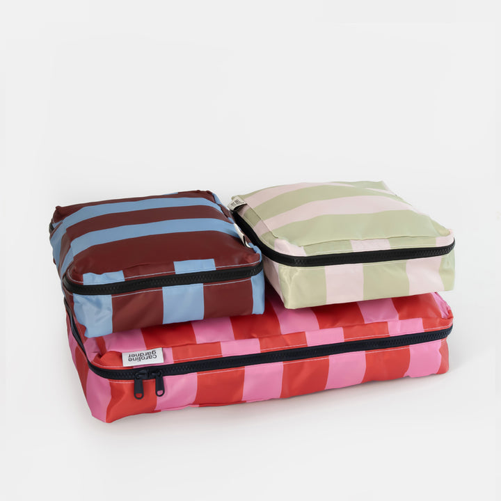 Three striped packing cubes stacked together on a light gray background. The bottom cube has pink and red stripes, the middle cube has brown and light blue stripes, and the top cube has pale green and white stripes.