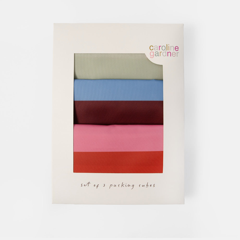 Caroline Gardner set of 3 packing cubes in a cream-colored cardboard box with a clear window showing the stacked cubes in sage green, light blue, burgundy, pink, and terracotta.