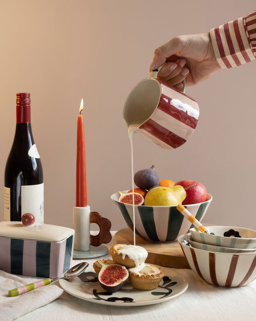 A hand in a red and white striped shirt pours a white liquid from a matching striped ceramic pitcher into a bowl of fruit. The scene is styled with a wine bottle, a lit red candle, a striped tin, and other ceramic tableware on a table.