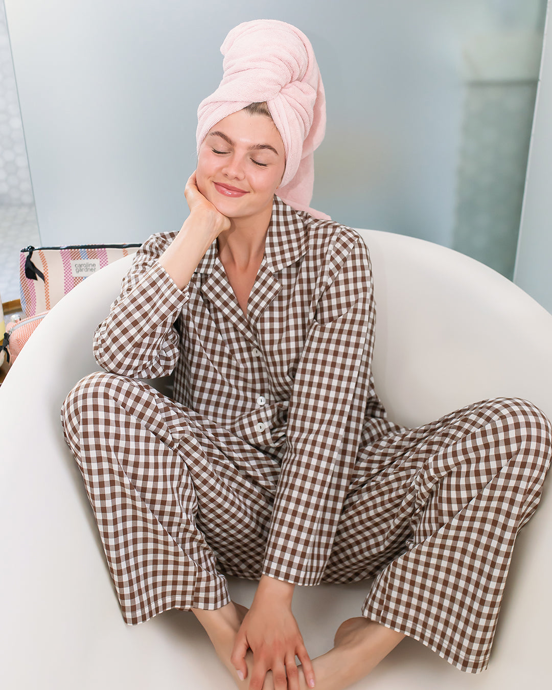A woman wearing a brown and beige gingham pajama set sits cross-legged on a white oval chair, with a pink towel wrapped around her hair. The background is a softly blurred indoor setting.