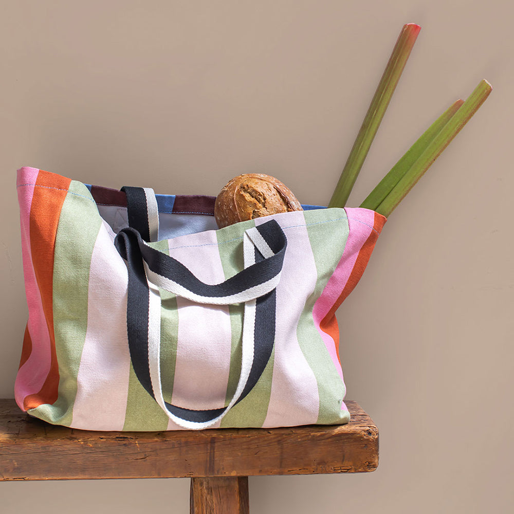 A striped canvas tote bag with dark navy handles sits on a rustic wooden table, styled with a loaf of bread and green plant stems against a plain beige background.