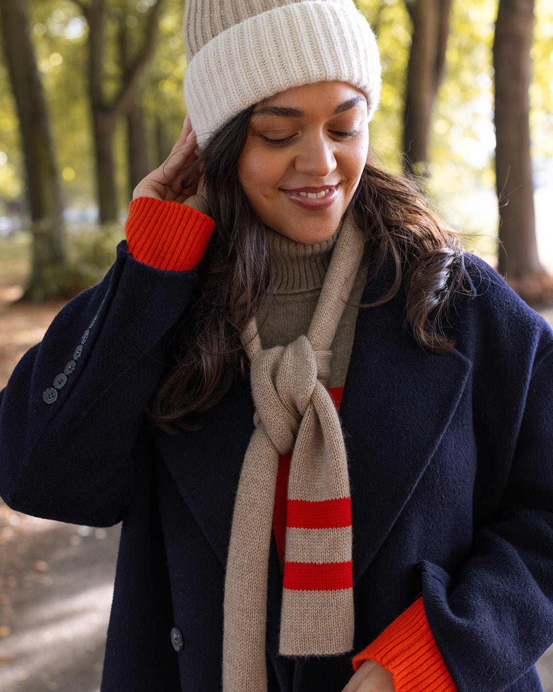 A woman outdoors wearing a cream knit beanie, a navy coat, a beige scarf with red stripes, and a red sweater, with trees in the background.