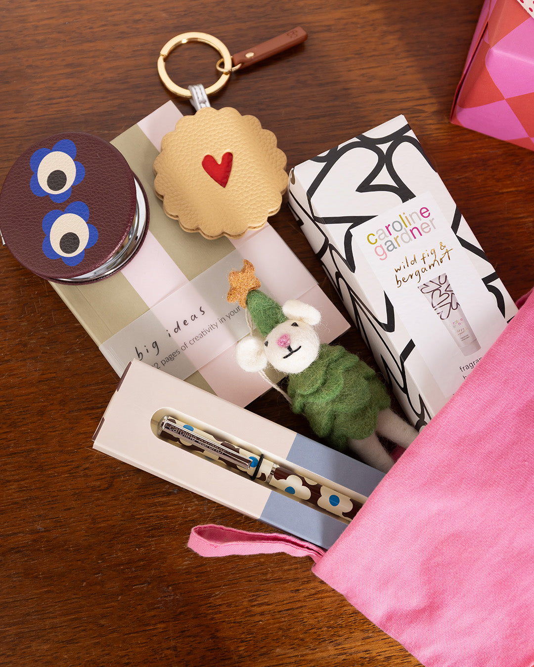 A styled flat-lay of gift items arranged on a wooden surface, including a pink drawstring pouch, a patterned box labeled 'caroline garavel', a small felt sheep character, a cookie-shaped keychain with a red heart, and other gift items.