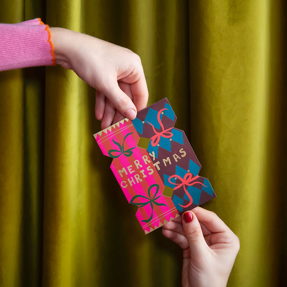 A person's hands holding a colorful Christmas card with 'MERRY CHRISTMAS' text, set against a draped olive green velvet curtain background.
