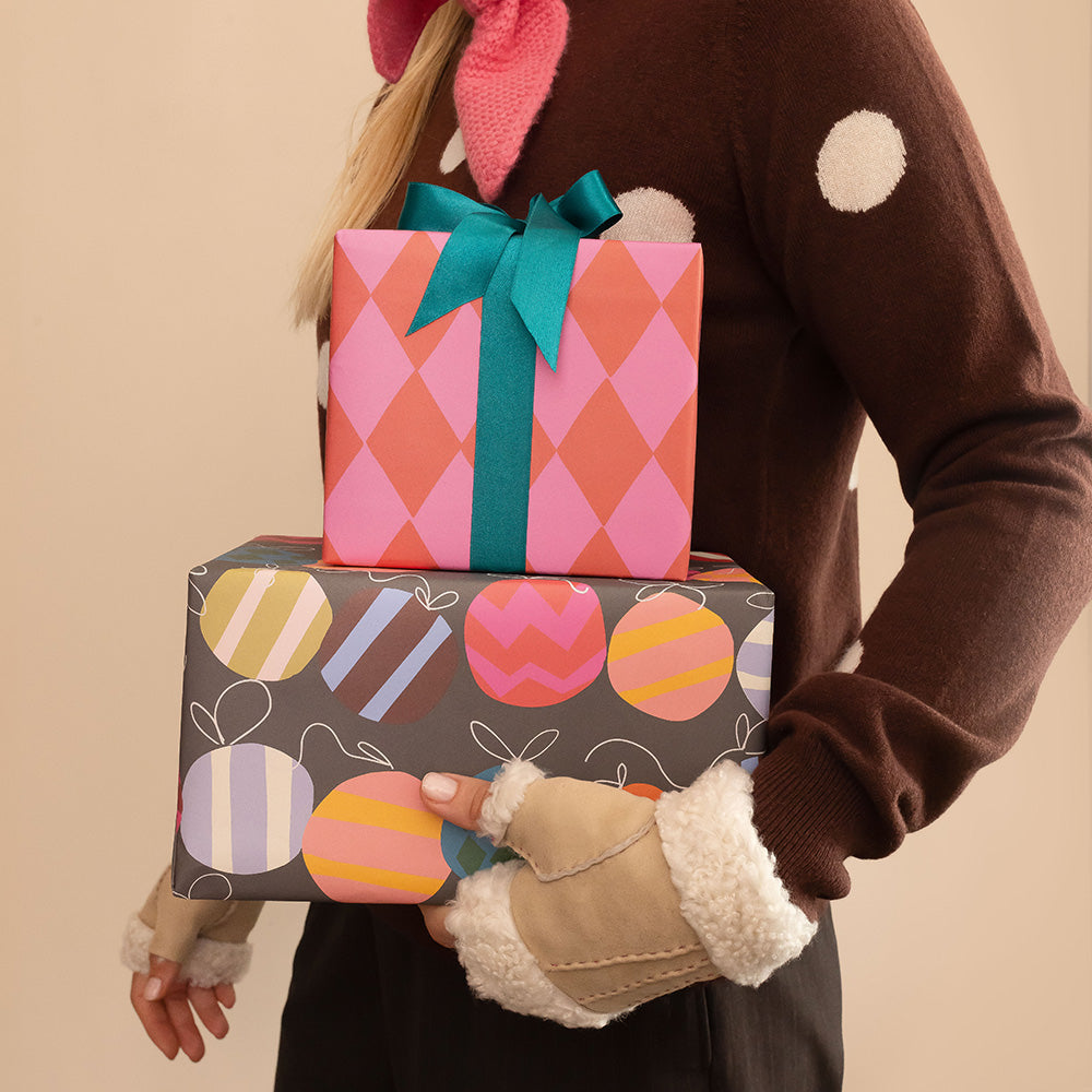 A person wearing a brown sweater and cream fleece fingerless gloves holds two wrapped gift boxes. The top box has pink and coral argyle wrapping with a teal bow, and the bottom box is dark gray with colorful striped and oval patterns.