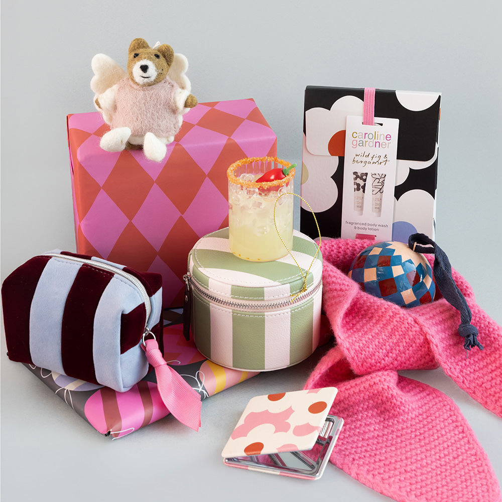 A curated gift set featuring a pink and orange argyle box with a plush teddy bear Christmas decoration, a striped cosmetic pouch, a round striped tin, a pink knit scarf, a patterned ball, a drink, and branded packaging on a light grey background.
