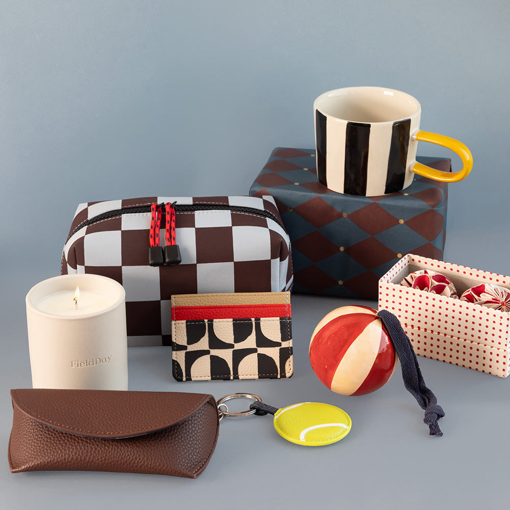 A styled flat-lay collection of various accessories and home items on a solid blue-gray background, including a brown and white checkered zippered pouch, a white candle, a brown pebbled leather pouch, a striped mug, a patterned card holder, a tennis ball keyring, and a small gift box.