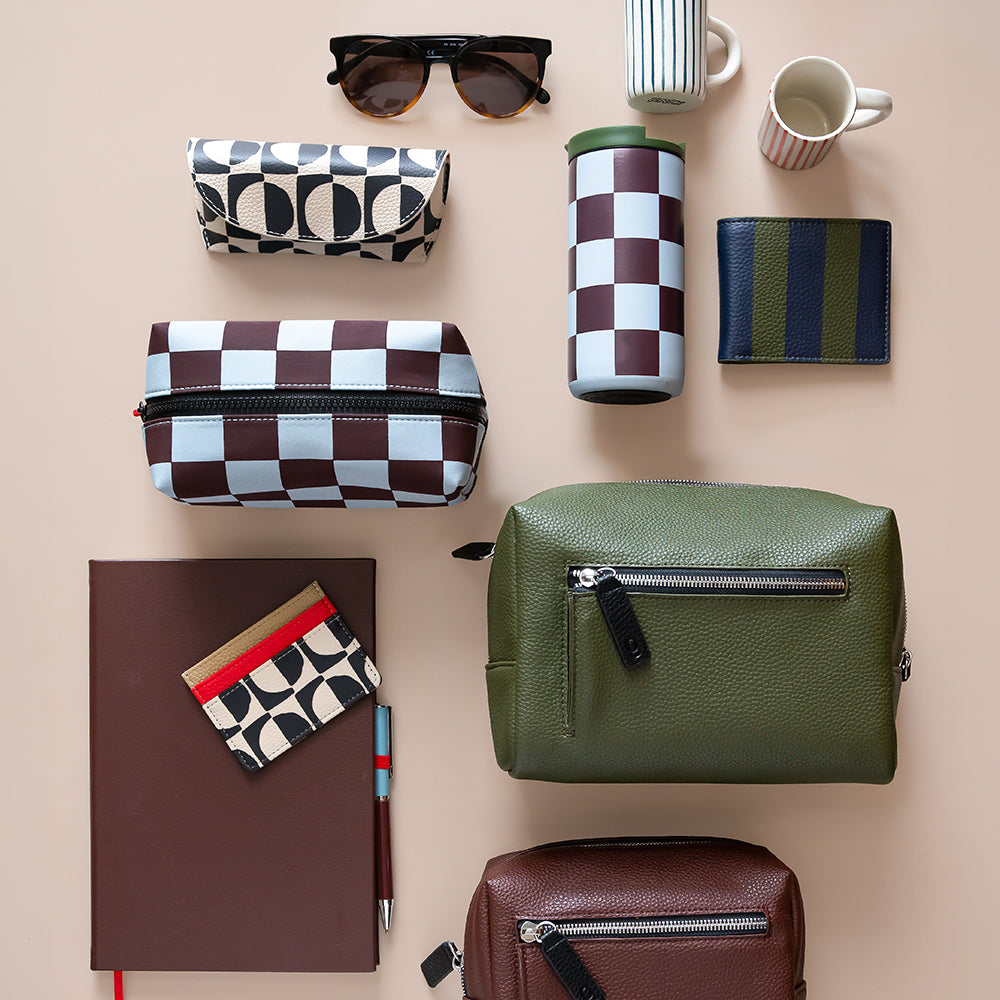 A styled flat-lay of multiple accessories and small goods on a warm beige background, including two checkerboard zip pouches, sunglasses, a striped tumbler, a striped wallet, a patterned card holder, a notebook, a pen, a mug, and two brown leather pouches.