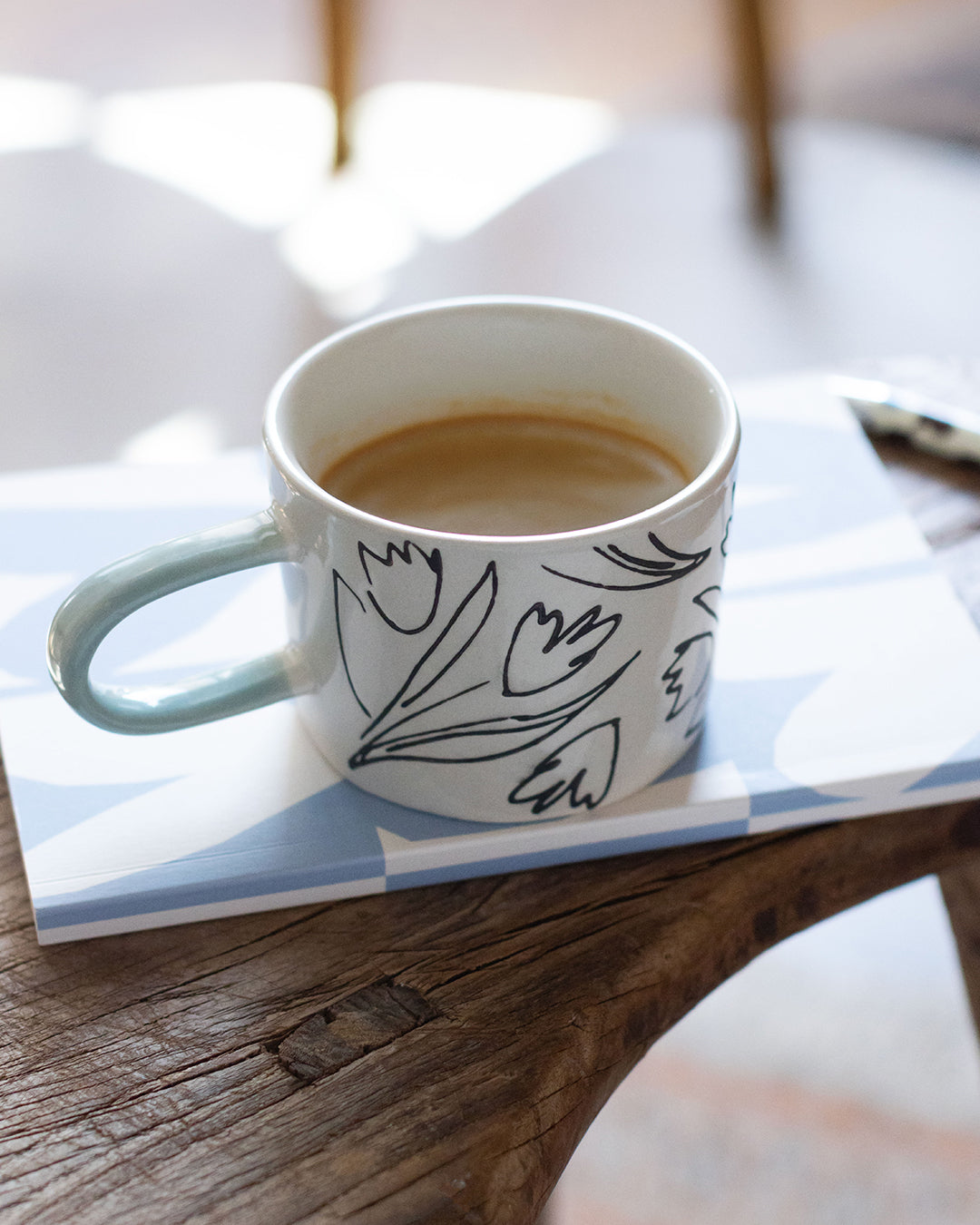 A white ceramic coffee mug with a black floral line drawing and a light blue-green handle, filled with coffee and resting on a blue-and-white patterned coaster on a rustic wooden table.