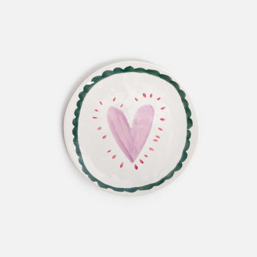 A round white ceramic plate with a scalloped dark green rim, featuring a hand-painted pink heart in the center surrounded by small red dots, set against a plain light gray background.