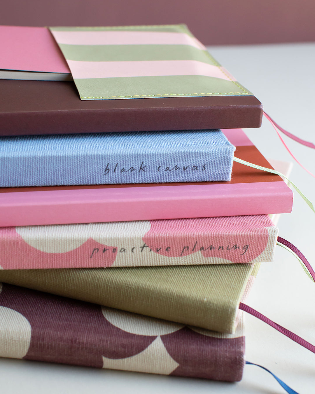 A stack of fabric-covered notebooks or journals with different color and patterned covers, including pink, blue, brown, and striped designs, arranged on a white surface.