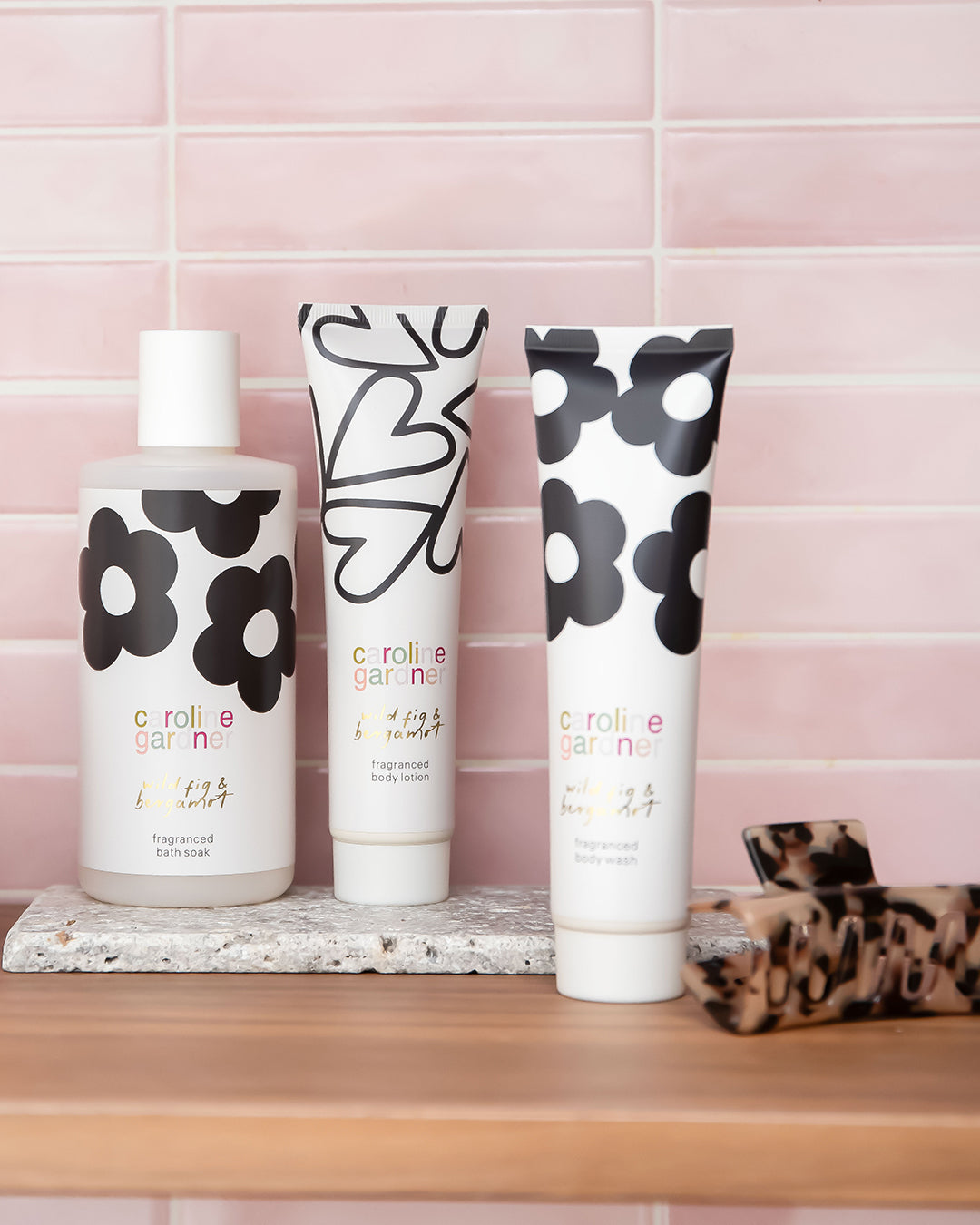 Three white toiletry products with black floral graphics and the brand name 'caroline gardner' are arranged on a wooden surface against a pink tiled background. A small leopard-print hair tie sits to the right.