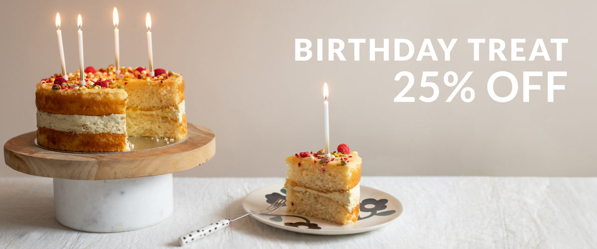 A birthday cake with lit candles on a wooden-and-marble stand, with a slice plated nearby and large text reading 'BIRTHDAY TREAT 25% OFF' on a beige background.