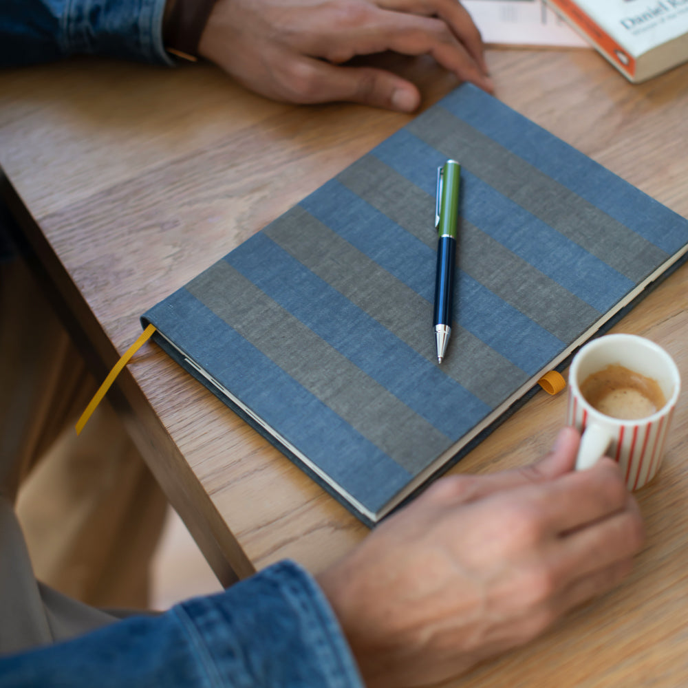 Person holding a coffee cup next to a blue notebook with a pen on a wooden table