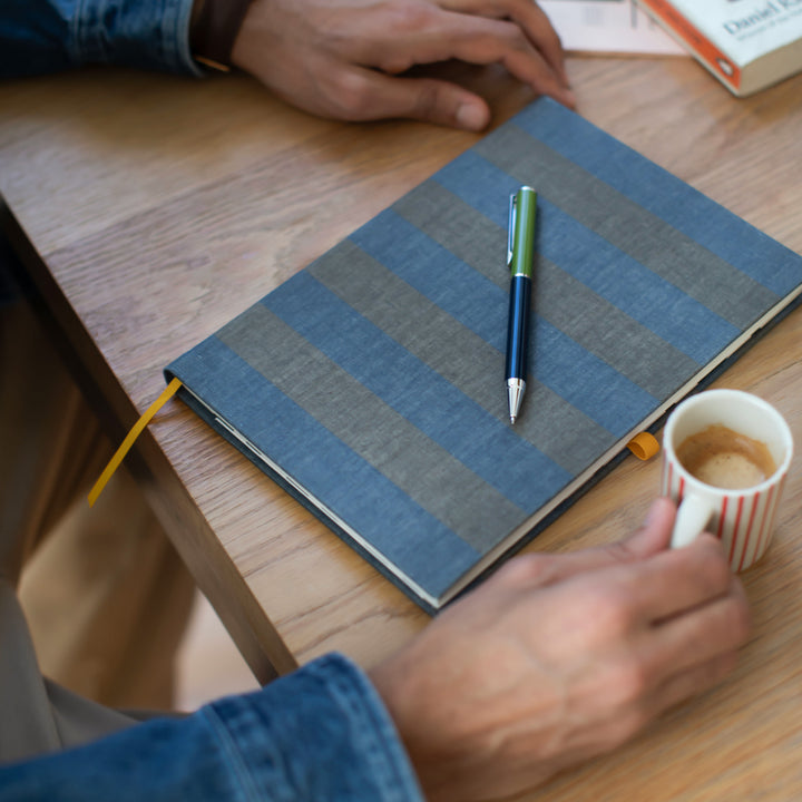 Person holding a coffee cup next to a blue notebook with a pen on a wooden table
