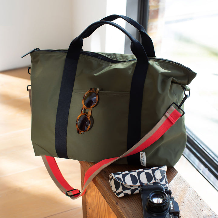 Green duffel bag with sunglasses and camera on a wooden surface near a window