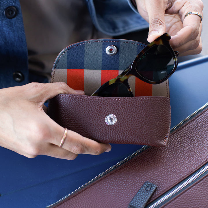 Person placing sunglasses into a checkered sunglasses case held over a blue leather bag.