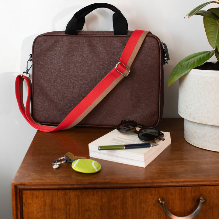 purple laptop bag with red strap on a wooden surface with books, sunglasses, pen, and keys.