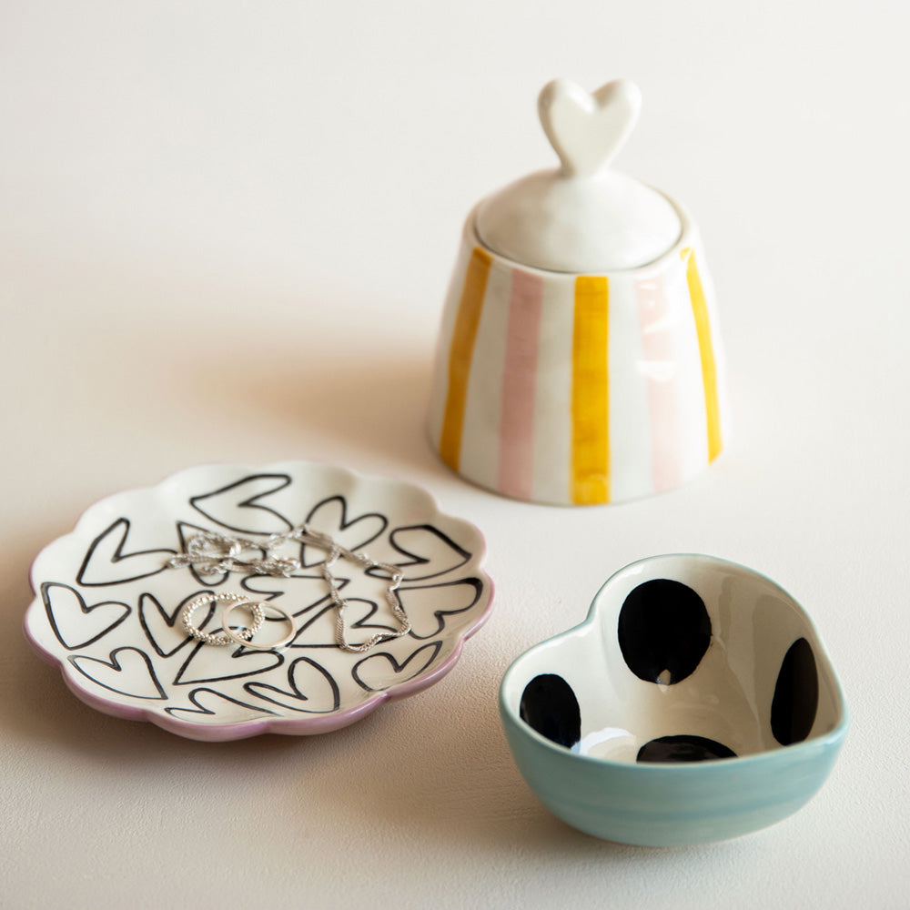 Three ceramic items including a heart-shaped dish, a striped storage pot with a heart lid, and a polka dot bowl on a light background.