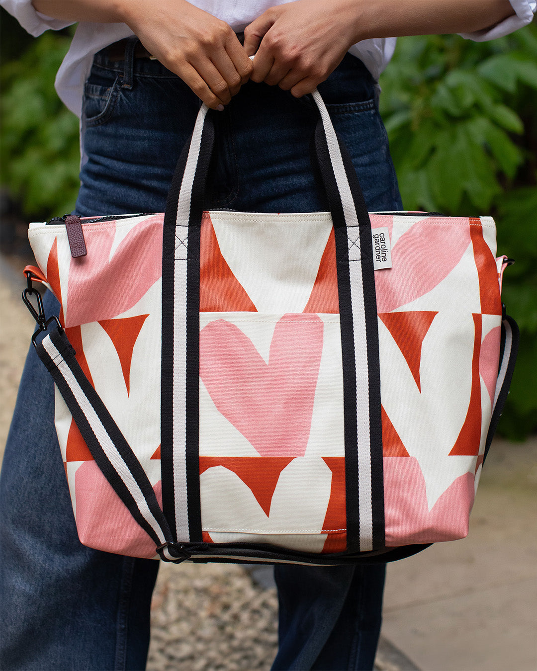 A person outdoors holding a large tote bag with a pink, white, and orange geometric heart pattern. The bag has black and white striped handles and a black-and-white shoulder strap.