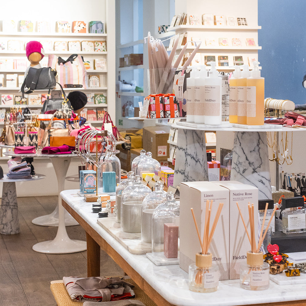 A retail display table in a boutique showcasing reed diffusers, glass jars, and boxed fragrance products, with shelves and other merchandise visible in the background.