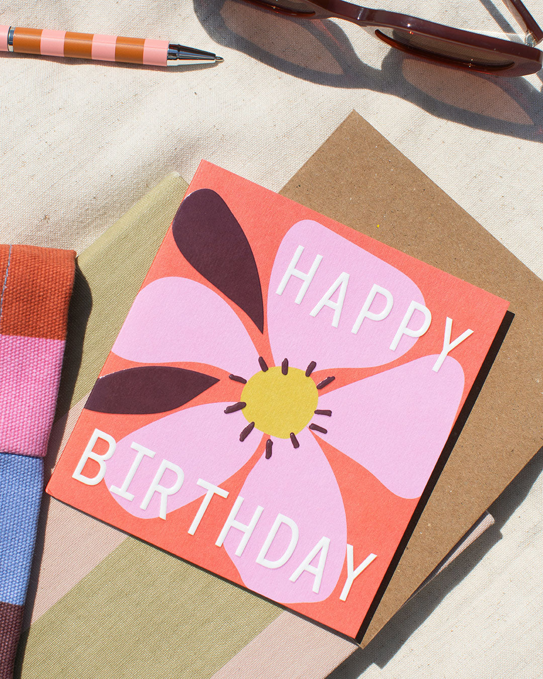 a pink and red birthday card with floral design, on a material surface with a pen and sunglasses