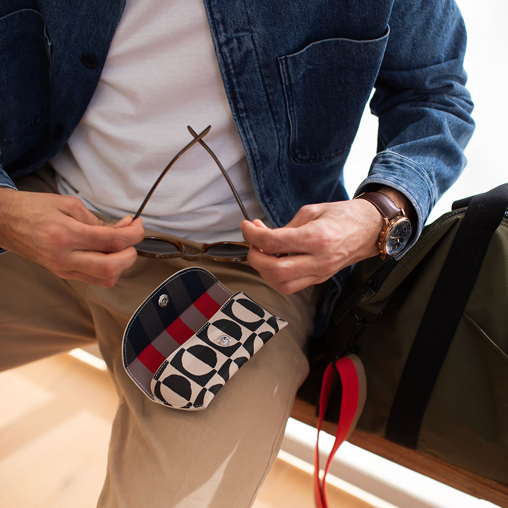 Person holding a patterned pouch with sunglasses, wearing a blue jacket and beige pants.