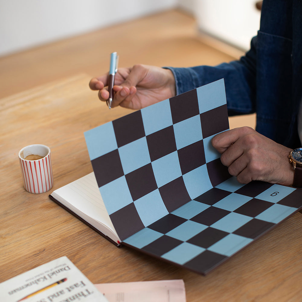 Person holding a checkered notebook with a pen, coffee cup, and book on a wooden table.